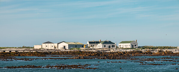 Remote white buildings on a rocky island shore with ocean water in foreground under clear blue sky for travel background