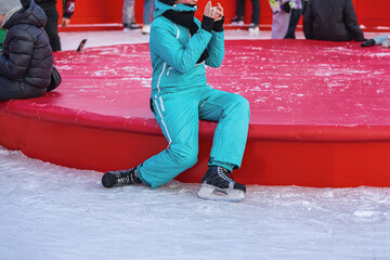 Girl making selfie photo on smartphone on ice rink. Person in turquoise snowsuit resting on red rink edge with ice skates on and taking photo on phone. Winter fun, ice skating break, outdoor activity