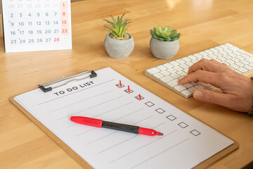 Hand typing on white keyboard at wooden desk beside clipboard with checked to-do list, red marker, calendar and small plants — organized workspace for planning and productivity
