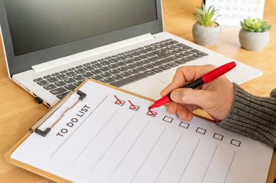 Hand with red marker checking off completed tasks on a to do list clipboard, symbolizing productivity, organization, planning, and goal achievement on a wooden desk with a laptop