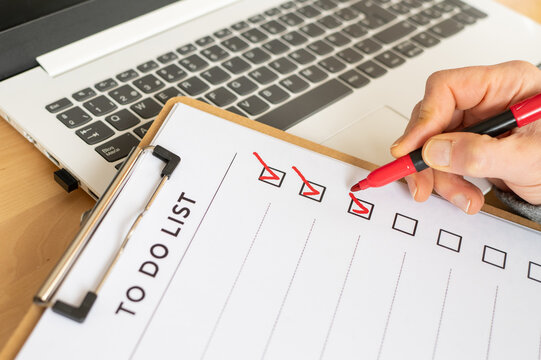 Hand holding a red marker pen checking off completed tasks on a to do list document clipped to a clipboard, with a laptop keyboard in the background, signifying task management and productivity