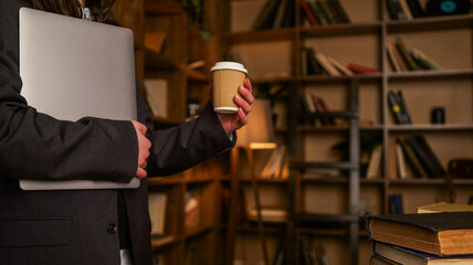 Professional holding a laptop and coffee cup in a library with bookshelves © shine.graphics