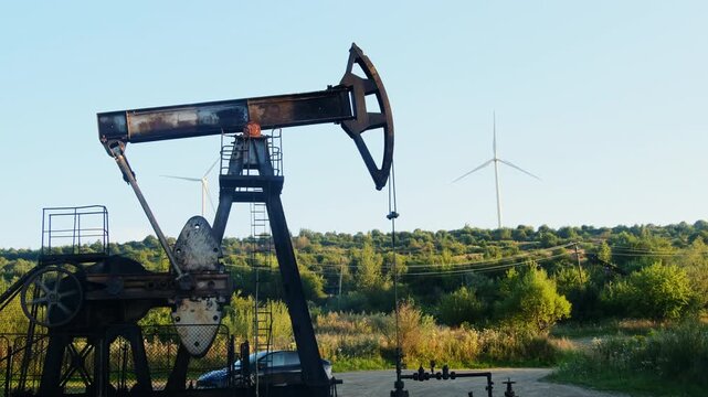 Rusty pumpjack stands on field while wind turbine turns beyond trees. Walking beam dips near horsehead lifting with counterweight rotating steadily