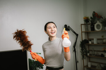 An Asian woman wearing orange rubber gloves smiles while holding a feather duster and spray bottle...
