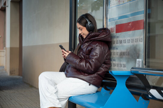 Teen girl smiling while looking at smartphone and listening to music at bus stop, casual winter clothes, medium side view, relaxing moment in urban public space
