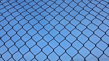 Perspective view of a metal wire mesh fence or chain link fence against a clear blue sky background. Concept of security, boundary, or restriction.