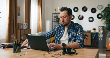 Fototapeta premium A serious bearded man wearing casual clothes works online as a support operator. He concentrates on helping customers, solving problems, and responding to digital inquiries from home.