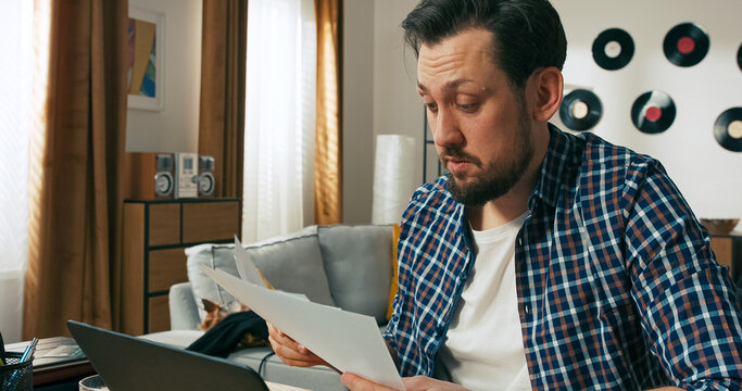 A tired man with a beard looks down at unpaid bills, visibly overwhelmed. His focus is intense as he searches for ways to manage money problems and settle his financial obligations.