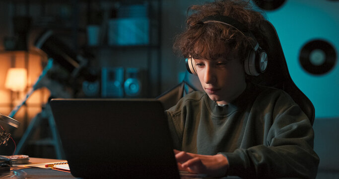 Smiling young college-aged boy has wireless headphones listens to music and works at computer, writes paper for school at desk at home.