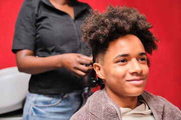 Afro young man smiling in salon as hairstylist braids his hair from behind