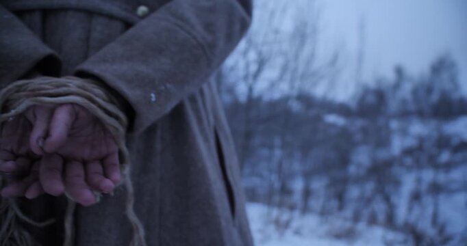 Cinematic close up of a prisoner's hands tied firmly with a rope behind their back while wearing a vintage wool coat in a freezing snowy winter landscape.