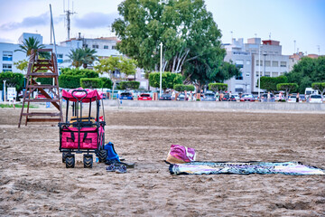 Beach setup with cart, towel, and shoes near lifeguard tower and parked cars during daytime