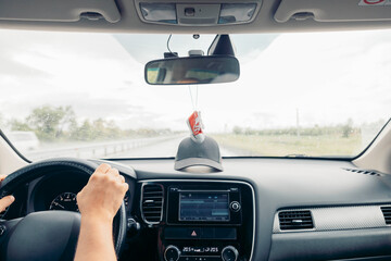 driver holds steering in car on rainy road, windshield wipers working, dashboard screen visible, front view of travel journey with mirror and hanging decoration, transport, lifestyle