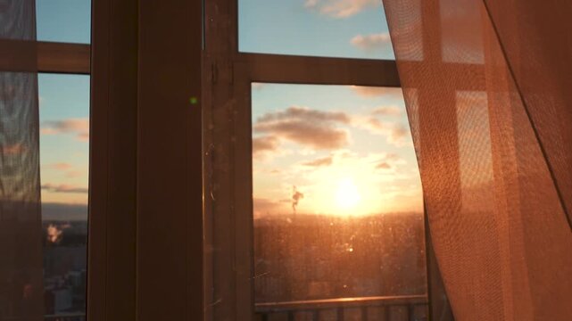 Human hand gently opening transparent sheer curtain to reveal warm sunset over city with golden sunbeams shining into room. Symbolizing hope curiosity and new beginning.