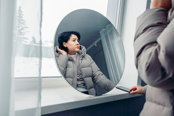 A sad Asian woman with short black hair is fixing her short black hair while looking in a round mirror above her wrinkles. It is snowing outside, indicating that it is winter time.