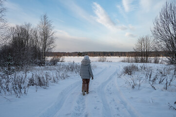 A woman in winter clothes walks along a snow-covered road in the forest on the bank of a frozen river.
