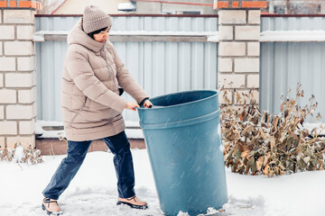 middle-aged Asian woman in a beige coat and hat pushes a large blue trash bin through the snow in a winter setting.