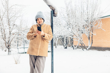 young Asian woman with smartphone in a snowy landscape. She wears a beige coat and a gray beanie. Snow-covered trees and a wooden house are in the background