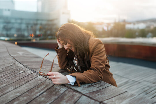 Joyful woman in brown jacket smiling while resting on wooden bench, natural light, urban background, concept of carefree moment and happiness
