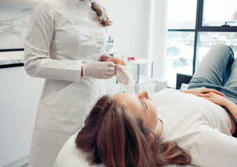 A female cosmetologist in a sterile environment performs a skin care treatment on a young woman. The doctor wears gloves and a white coat, ensuring cleanliness.