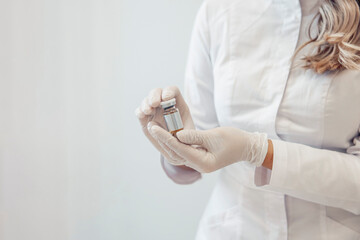 A female cosmetologist in a white coat holds a vial of serum. She wears gloves and has long, wavy hair. The setting is clean and sterile, focused on skin care.