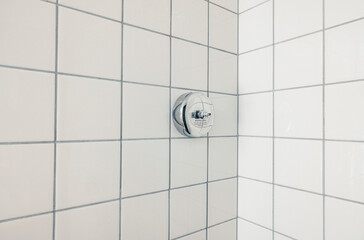 A close-up view of a bathroom corner with white ceramic tiles. The tiles are arranged in a grid pattern, featuring a chrome shower control in the center.