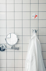 A modern bathroom with white ceramic tiles. A round mirror is mounted on the wall. A white bathrobe hangs on a hook, adding a touch of comfort.