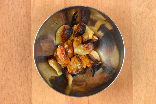 A top down view shows several golden brown roasted garlic cloves with charred edges served in a small round metal bowl on a light wooden background.