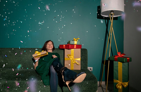 Happy woman sitting on sofa with gifts and confetti in festive living room