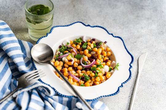 Close-up of a bowl of chickpea, red onion, pomegranate and coriander salad with a glass of mint water on a table