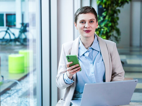 Businessperson with smartphone and laptop at modern office looking at camera