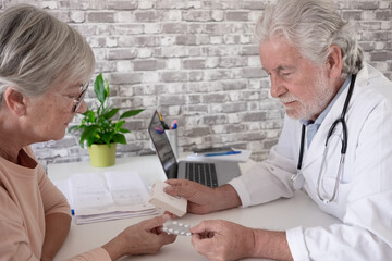 Senior male doctor consulting with elderly female patient in a medical office, reviewing documents at a desk with a laptop. Concept of healthcare, medical consultation, diagnosis, trust, patient care