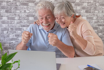 Happy elderly couple laughing expressing joy upon discovering a lottery win, two modern seniors using laptop at home enjoying technology