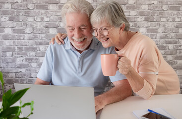 Happy senior couple using a laptop together at home, looking at the screen while enjoying a coffee cup. Concept of active aging, digital literacy, online banking, communication, technology
