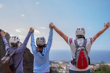 Rear view of carefree active group of seniors with raised arms hiking on mountain wearing protective helmets looking at horizon over sea.