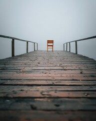 Fototapeta premium A solitary orange chair at the end of a weathered wooden pier, fading into a foggy gray horizon. Concept Solitary Chair, Weathered Pier, Foggy Horizon, Orange Accent, Moody Seascape