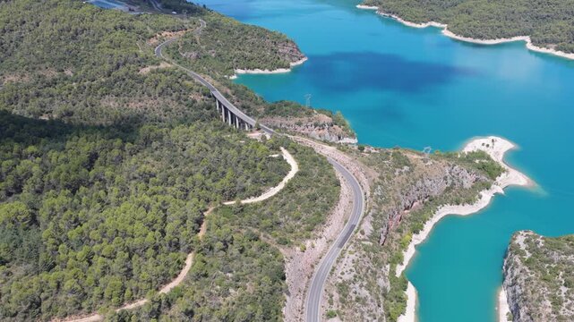 Aerial view of a bridge spanning a turquoise lake surrounded by lush green forests and winding roads, creating a striking contrast of colors, El Grado, Aragon, Spain.