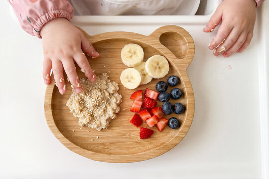 Caucasian baby learning self feeding with oatmeal, banana and berries on wooden plate. Baby led weaning concept, healthy toddler nutrition and safe home mealtime routine.