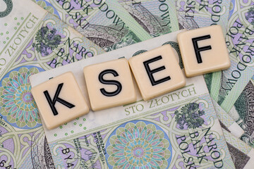 Blocks with the abbreviation KSEF, standing for National E-invoice System, are placed against a background of loosely spread Polish 100 PLN banknotes. The photo was taken under artificial lighting.