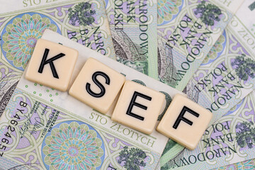 Blocks with the abbreviation KSEF, standing for National E-invoice System, are placed against a background of loosely spread Polish 100 PLN banknotes. The photo was taken under artificial lighting.