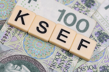 Blocks with the abbreviation KSEF, standing for National E-invoice System, are placed against a background of loosely spread Polish 100 PLN banknotes. The photo was taken under artificial lighting.