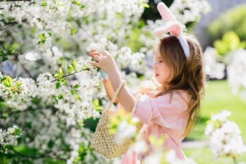 Easter egg hunt. Girl child Wearing Bunny Ears Running To Pick Up Egg In Garden. Easter tradition. Baby with basket full of colorful eggs.