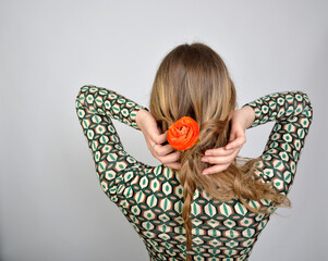 Detailaufnahme einer Person mit blondem Haar, das zu einer kunstvollen Form gebunden ist, in die eine leuchtend orangefarbene Blume integriert wurde. Die Hände arrangieren sorgfältig die Haare und set