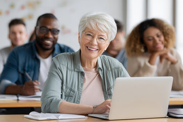 Smiling senior woman with glasses using a laptop in a diverse classroom setting for lifelong learning education technology and modern retirement lifestyle concepts