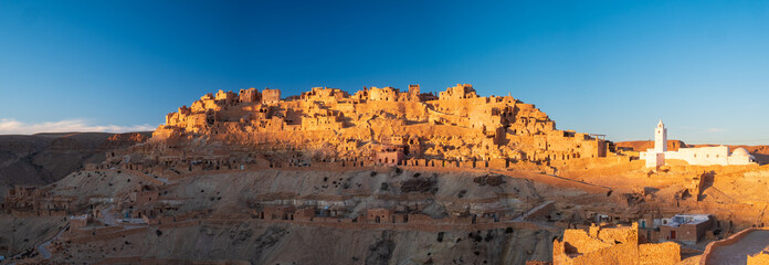 Chenini ksar at golden hour in southern Tunisia, ancient Berber village with white mosque and stone houses on desert hill, dramatic light, heritage, travel destination
