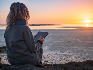 Woman using tablet on quiet beach in winter at sunset, back view, calm seascape in Djerba Tunisia, remote work, solitude, travel lifestyle