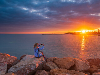 Woman photographing dramatic sunset over calm sea from rocky shore in Djerba Tunisia, travel photography, golden light, peaceful moment, tourism