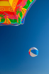 Vibrant detail of a colorful beach umbrella and inflatable beach ball contrasting against a deep blue sky, capturing the essence of sunny summer fun and relaxation.