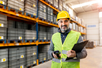 Smiling warehouse manager in safety vest and hardhat holding clipboard and scanner in a large distribution center with industrial shelves.