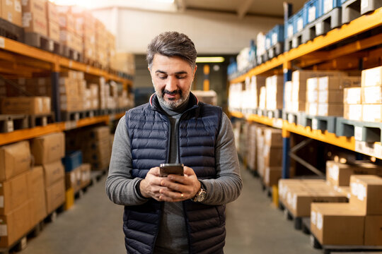 Middle-aged warehouse manager using smartphone in storage. Professional businessman in distribution center. E-commerce, supply chain and technology concept. Logistics worker.